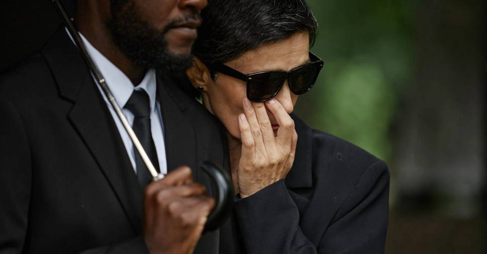 Homem de terno apoia mulher enlutada sob guarda-chuva em funeral de emergência no fim de semana.